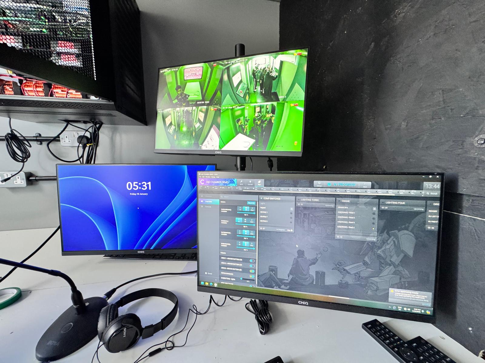 The facilitation and control desk outside The Chamber, with headphones, a microphone, a monitor showing four live camera feeds and a wide screen running the Chamber control system.