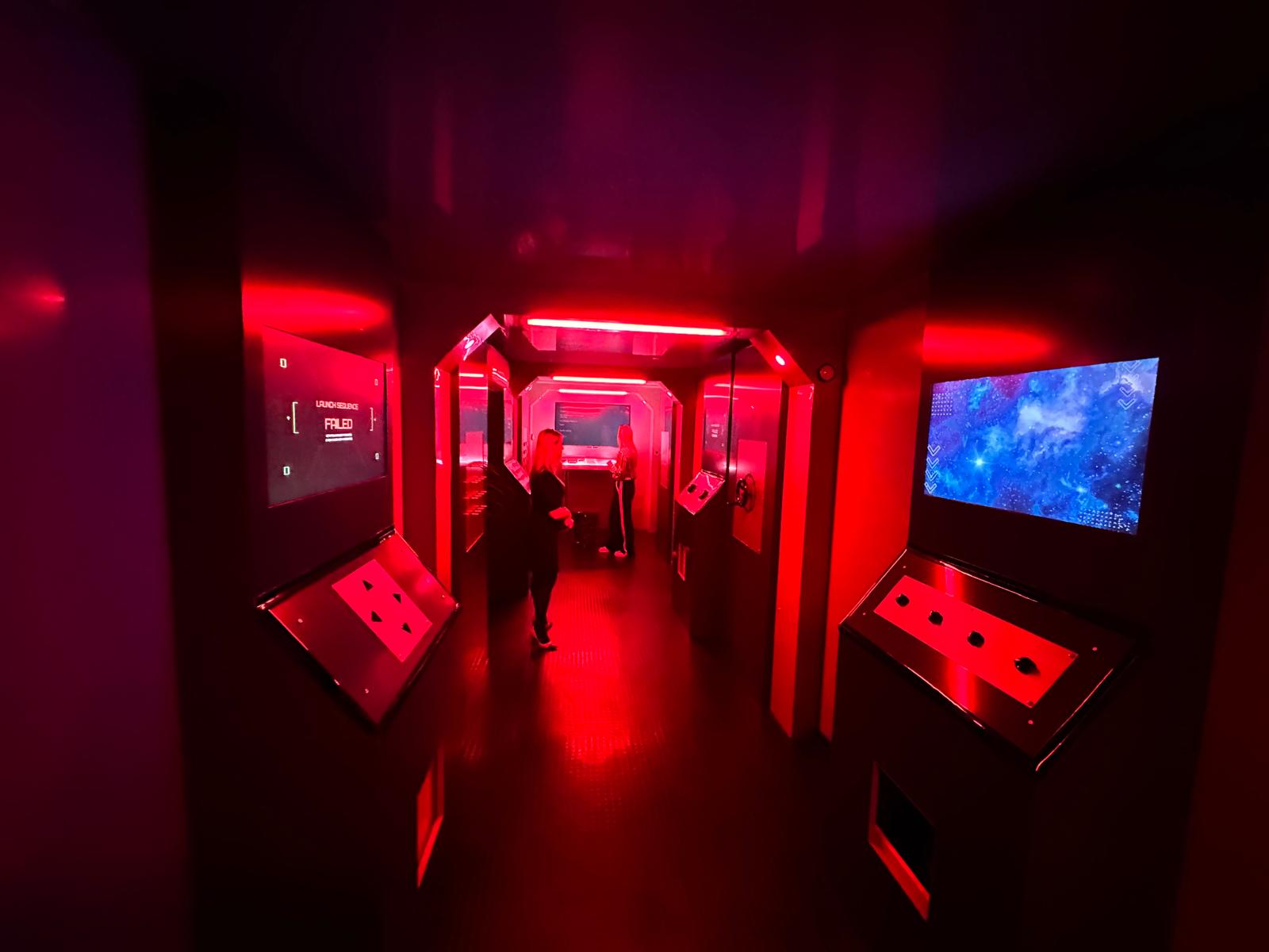 The Chamber corridor under deep red lighting mid-scenario, with two participants between angled control consoles, a wall screen reading launch sequence failed and lit arches leading deeper into the environment.