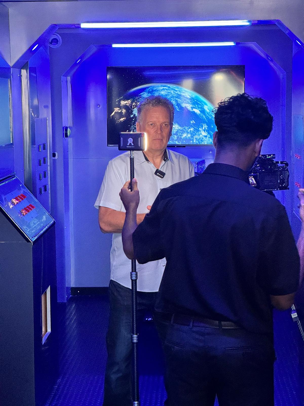 A presenter being filmed inside The Chamber under blue lighting, with a camera operator, boom light and a screen showing Earth from orbit behind him.
